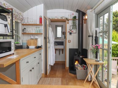 A small kitchen area with wooden countertops and cabinets a wood stove with fire a sink through a doorway and a small table with flowers near glass doors at Burr 2 Shepherds Hut Swimbridge near Barnstaple