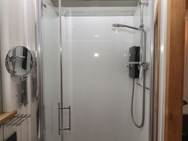 A shower enclosure with a glass door including a showerhead soap dispenser and a wall-mounted mirror in a bathroom at Burr 2 Shepherds Hut in Swimbridge near Barnstaple