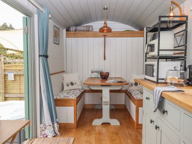 A dining area with a wooden table and bench seats with cushions inside a hut with a kitchen counter and appliances on the right at Burr 2 Shepherds Hut Swimbridge near Barnstaple
