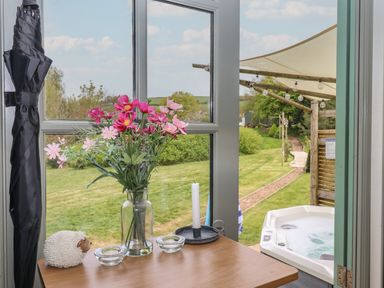 A table with flowers, a candle, and a sheep ornament by a window overlooking a garden and a hot tub at Burr 2 Shepherds Hut Swimbridge near Barnstaple