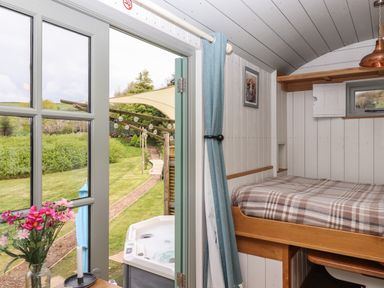 A bedroom with a bed and a small window beside an open door leading to a garden with a hot tub and pathway at Burr 2 Shepherds Hut in Swimbridge near Barnstaple