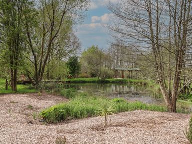 A small pond surrounded by trees and grassy areas with a wooden dock and a covered seating area at Burr 2 Shepherds Hut in Swimbridge near Barnstaple