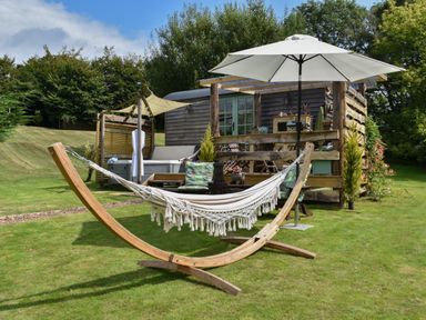 An outdoor area with a hammock on a wooden stand and a white umbrella near a wooden hut with green doors at Burr 2 Shepherds Hut Swimbridge near Barnstaple