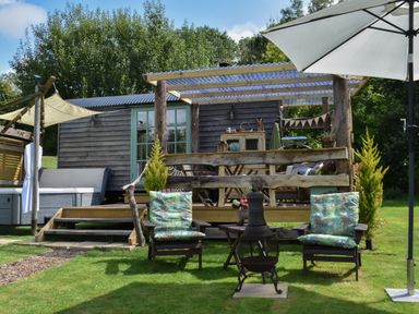 A wooden cabin with a covered porch two chairs with cushions a small table and fire pit on grass with an umbrella at Burr 2 Shepherds Hut Swimbridge near Barnstaple