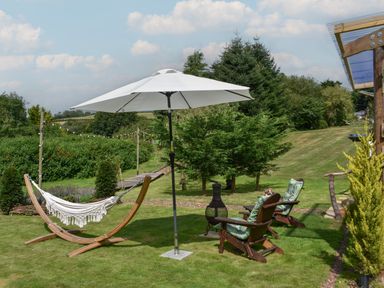 A garden with a hammock on a wooden frame a white umbrella two wooden chairs with cushions and green trees in the background at Burr 2 Shepherds Hut Swimbridge near Barnstaple