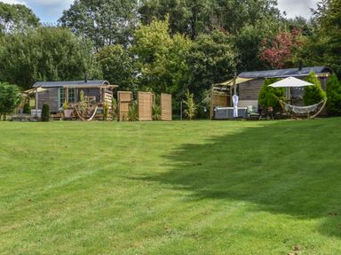 Two wooden shepherds huts with outdoor seating and hammocks on a grass lawn surrounded by trees at Burr 2 Shepherds Hut Swimbridge near Barnstaple