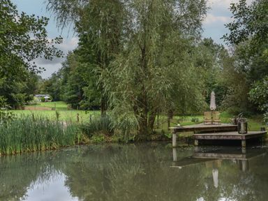 A pond with two wooden decks chairs and umbrella surrounded by trees and grass at Burr 2 Shepherds Hut in Swimbridge near Barnstaple