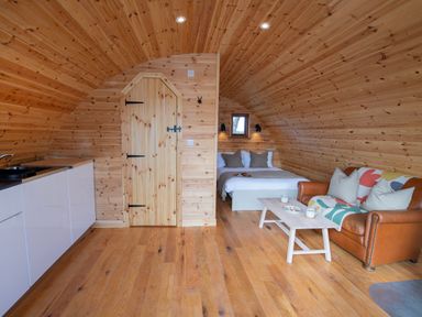 An interior wooden pod with a bed, a brown leather sofa, a coffee table, and a kitchen area with cabinets at Glamping Pod 1 Comfort in Lebberston