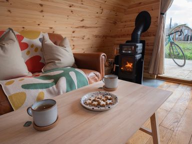 A wooden interior with a leather sofa and cushions a wood burning stove a table with two cups of tea and a plate of cookies a bicycle outside Glamping Pod 1 Comfort Lebberston