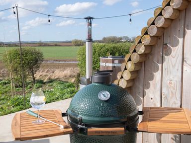 An outdoor Big Green Egg grill with wooden side tables a glass with a drink and a tool on one table and a wooden building with cut logs on the roof in a rural setting at Glamping Pod 1 Comfort in Lebberston
