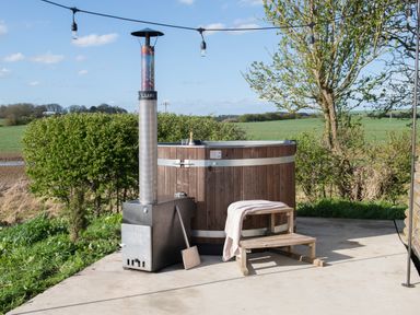 An outdoor hot tub with a wood stove heater on a concrete patio with a wooden step stool and string lights near trees and fields at Glamping Pod 1 Comfort in Lebberston