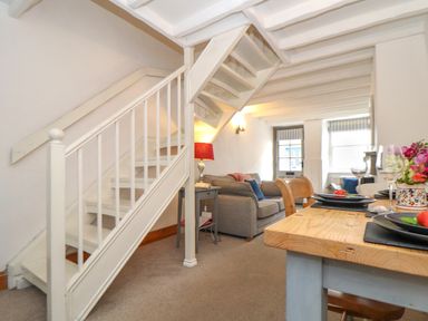 A living room with a staircase, gray sofa, side table with lamp, dining table set with plates and glasses at Cobblestone Cottage in Bude