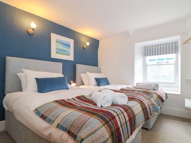A bedroom with two single beds with plaid blankets and rolled towels against a blue and white wall at Cobblestone Cottage in Bude