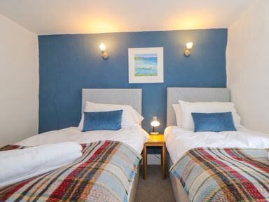 A bedroom with two single beds with blue cushions and plaid blankets and a wooden nightstand with a lamp against a blue accent wall at Cobblestone Cottage in Bude