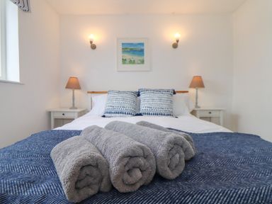A bedroom with a bed with three rolled towels on a blue blanket two patterned pillows two bedside tables with lamps a framed picture and two wall lights at Cobblestone Cottage in Bude