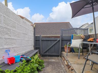 A small outdoor patio with a wooden bench holding children's beach toys and a seating area with chairs a table and an umbrella at Cobblestone Cottage in Bude