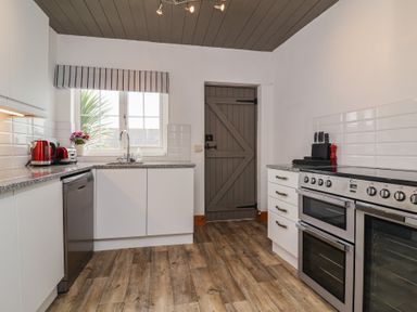 A kitchen with white cabinets, grey door, and stainless steel appliances at Cobblestone Cottage in Bude
