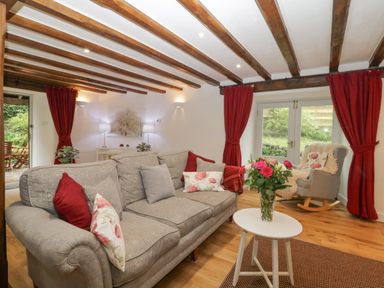 A living room with a grey sofa, red curtains, a rocking chair, and a small white table with flowers at 4 Barton Cottages Down Thomas near Plymouth