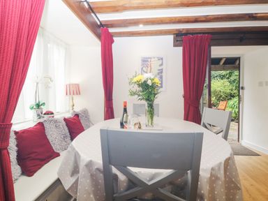 A dining area with a round table covered with a polka dot tablecloth surrounded by chairs and bench seating with cushions and red curtains at 4 Barton Cottages in Down Thomas near Plymouth