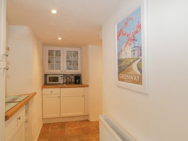 A narrow kitchen area with white cabinets a microwave a coffee maker glassware in a cabinet and a greenway poster on the wall at 4 Barton Cottages Down Thomas near Plymouth