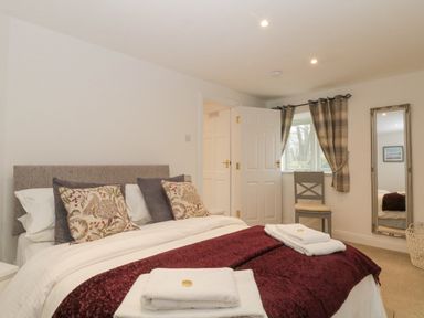 A bedroom with a bed with pillows and folded towels a chair under a window with curtains a wall mirror and an open door at 4 Barton Cottages in Down Thomas near Plymouth