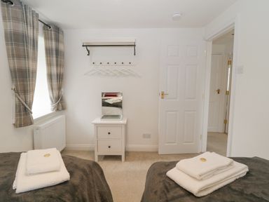 A bedroom with two beds each with folded towels on top a white nightstand with a mirror on it plaid curtains on the windows at 4 Barton Cottages in Down Thomas near Plymouth