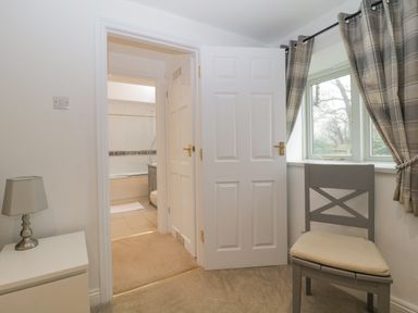 A room with a gray chair by a window with curtains and a white bedside table with a lamp next to an open door leading to a bathroom at 4 Barton Cottages Down Thomas near Plymouth