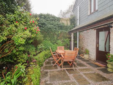 A patio area with wooden table and chairs surrounded by plants and flowers at 4 Barton Cottages in Down Thomas near Plymouth