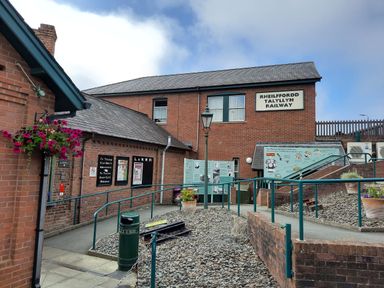 An exterior view of a brick railway building with signs and a ramp leading to the entrance at Rheilffordd Talyllyn Railway