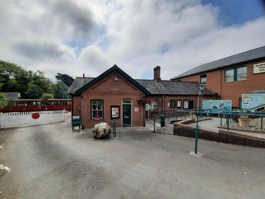 A train station platform with a brick building and a red train on the tracks at Hill View in Bryncrug near Tywyn