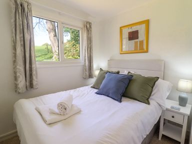 A bedroom with a bed with pillows and rolled towels by a window with curtains and a bedside table with a lamp at Hill View in Bryncrug near Tywyn