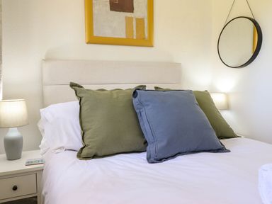 A bedroom with a bed with green and blue pillows a bedside table with a lamp a round wall mirror and a framed painting at Hill View in Bryncrug near Tywyn