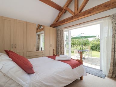 A bedroom with a bed white bedding and red pillows wooden wardrobes exposed wooden beams and a patio door leading to a garden with a table chairs and a green umbrella at Orchard View Redmary D Abitot near Hartpury