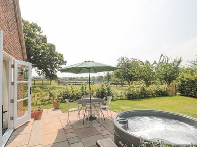 A patio with a table and chairs under a green umbrella and a hot tub next to a garden at Orchard View in Redmary D'Abitot near Hartpury