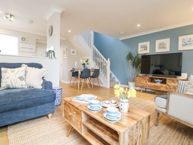 A living room with a wooden coffee table with cups and flowers and a large television on a wooden stand at Breeze Cottage in Falmouth