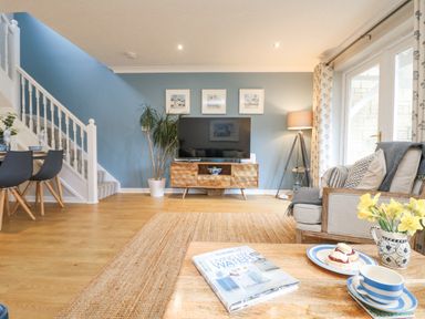A living room with a wooden coffee table, sofa, TV on a wooden stand, dining table with chairs, and staircase at Breeze Cottage in Falmouth