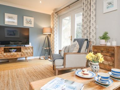 A living room with a wooden coffee table with cups and daffodils and an armchair next to a wooden cabinet and TV at Breeze Cottage in Falmouth