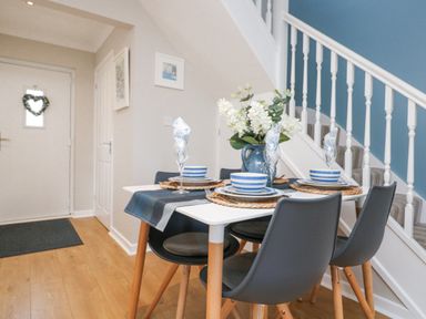 A dining area with a white table and four gray chairs set with plates bowls glasses and a vase with flowers near a staircase at Breeze Cottage in Falmouth