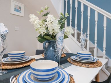 A dining table set with blue and white striped dishes glasses with patterned napkins and a blue vase with white flowers near a staircase at Breeze Cottage in Falmouth