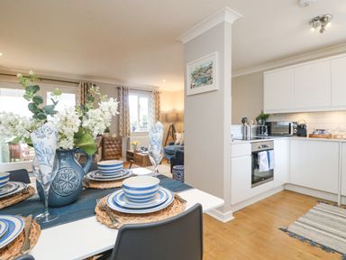 A dining table set with blue and white striped dishes and napkins with a vase of flowers next to a kitchen with white cabinets and wooden floor at Breeze Cottage in Falmouth