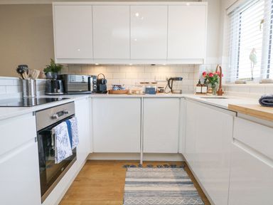 A kitchen with white cabinets a built-in oven a microwave coffee machine and a rug on the floor at Breeze Cottage in Falmouth