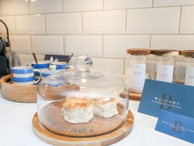 A kitchen countertop with scones under a glass dome a tray with striped cups and jars labeled sugar tea and coffee at Breeze Cottage in Falmouth