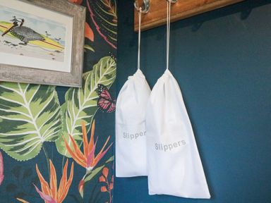 White bags labeled slippers hanging on hooks against a dark blue wall with tropical leaf and bird wallpaper at Breeze Cottage in Falmouth