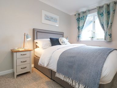 A bedroom with a bed a nightstand with a lamp and a window with curtains at Breeze Cottage in Falmouth
