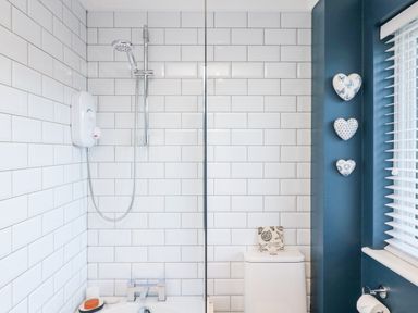 A bathroom with a shower, white tiled walls, a white toilet, and three decorative hearts on a blue wall at Breeze Cottage in Falmouth