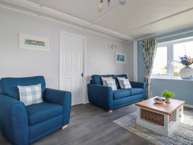 A living room with a blue armchair a blue sofa with pillows a wooden coffee table and a window with curtains at Lowen Lodge in St Merryn near Padstow