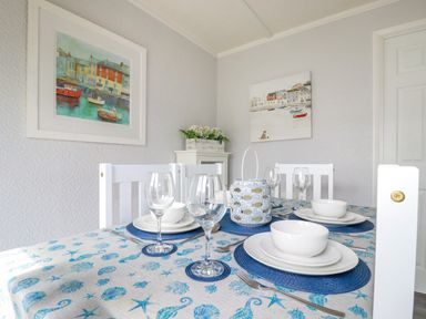 A dining table set with white plates bowls and wine glasses in a room with white chairs and coastal paintings at Lowen Lodge in St Merryn near Padstow