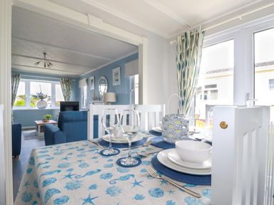 A dining table with blue and white tablecloth set with plates glasses and cutlery in a room with large windows and blue sofas at Lowen Lodge in St Merryn near Padstow