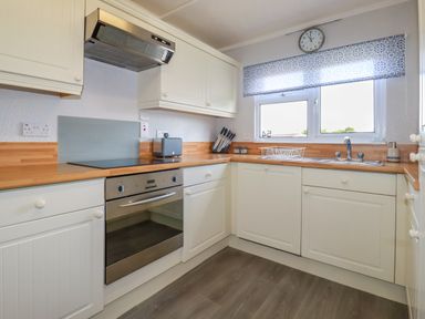 A kitchen with white cabinets wooden countertops a stainless steel oven stove a window with patterned blind and a clock above at Lowen Lodge in St Merryn near Padstow