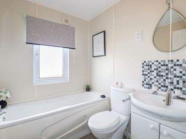 A bathroom with a bathtub under a window a toilet a sink with cabinet and a mirror at Lowen Lodge in St Merryn near Padstow